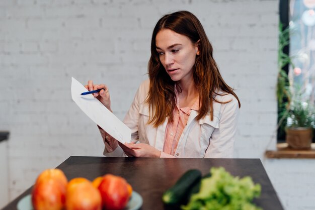 A person sitting at a desk, thoughtfully reviewing a printed meal plan with a pen in hand. The meal plan is detailed, including specific recipes and corresponding ingredients. A calendar is visible in the background, suggesting the integration of meal planning into the daily routine.