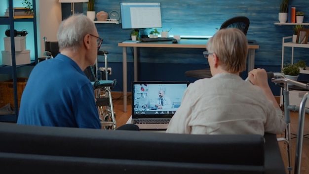 A doctor explaining a remote patient monitoring device to an elderly patient during a virtual consultation. The scene shows the practical application of telehealth technology, with the doctor pointing at the device on the patient's wrist while both are connected through a video call on a tablet.