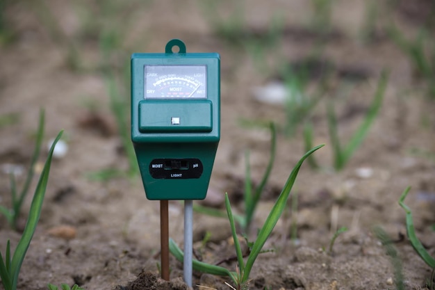 A close-up of an IoT sensor installed in the soil of a farm field. The sensor is connected to a small monitoring device, displaying real-time data on soil moisture and temperature. The background shows rows of crops.