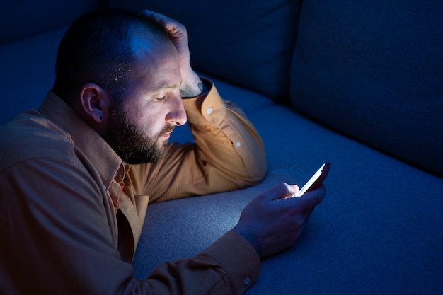 A person using a smartphone in bed late at night, with a blue light illuminating their face. The room is dark and the person looks tired.