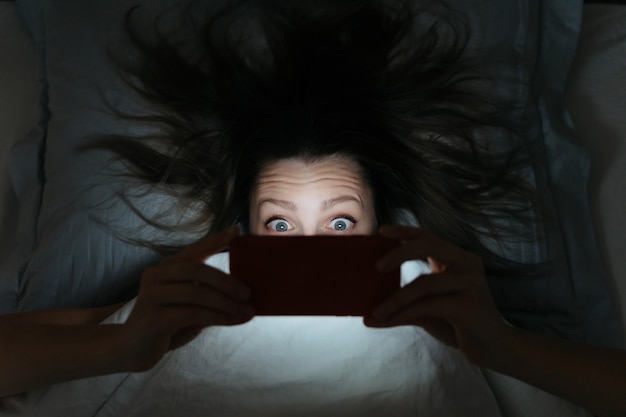 A person in bed, eyes wide open and unable to sleep, illuminated by the harsh light of a smartphone screen. The image captures the frustrating experience of insomnia caused by excessive screen time.